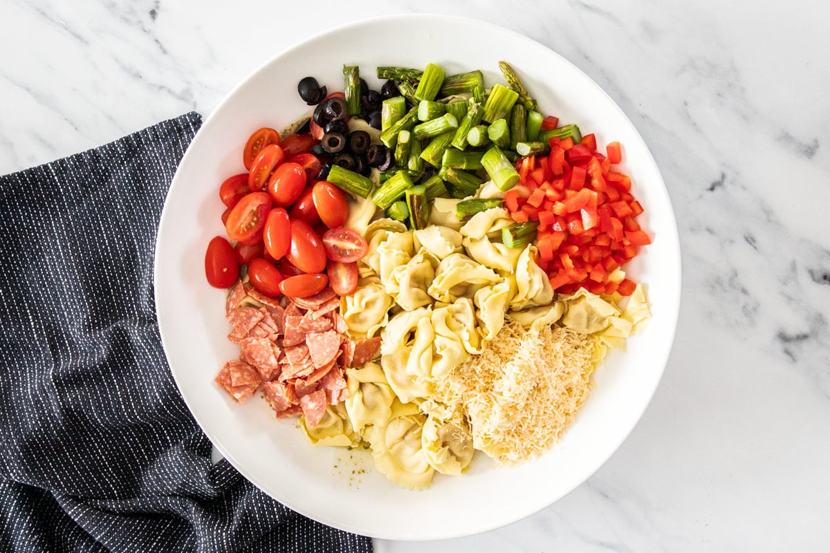 chopped peppers, asparagus, cheese, pepperoni, sliced olives and tomatoes and pasta in a white bowl.