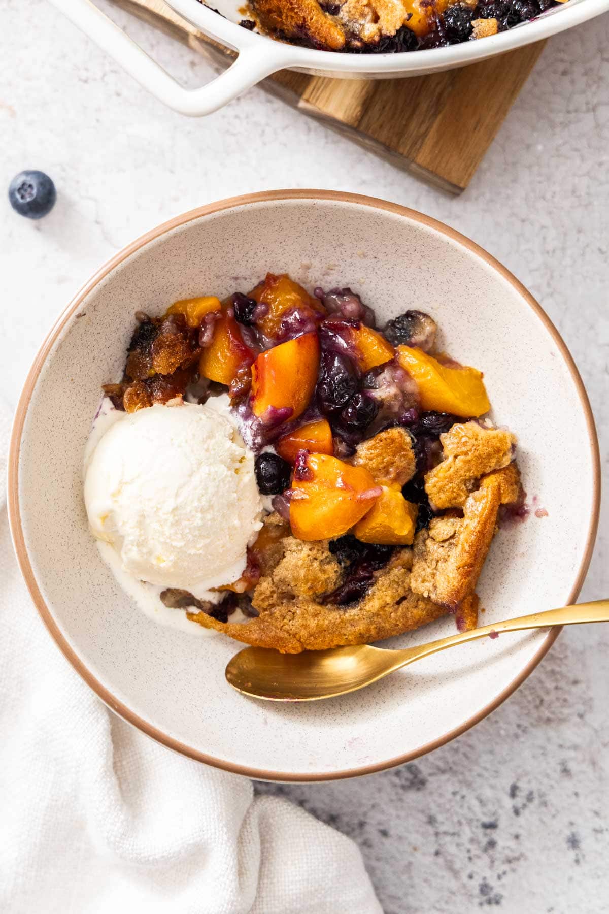 a bowl of blueberry and peach cobbler with a scoop of ice cream and a gold spoon.