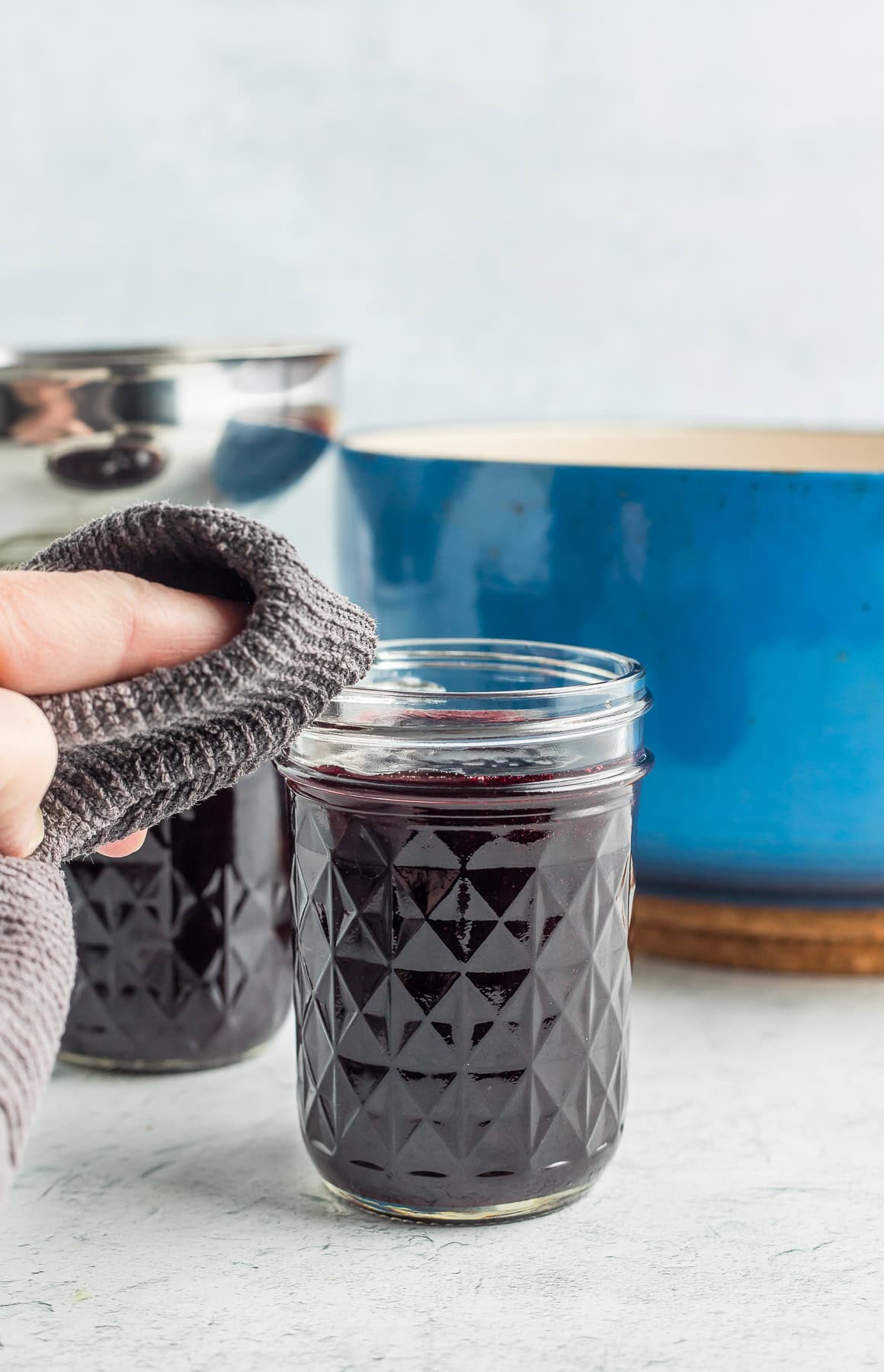 a hand using a grey towel to wipe a canning jar in front of a blue Dutch oven.