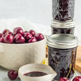 three jars and a small dish of cherry preserves with a white bowl of cherries in the background.