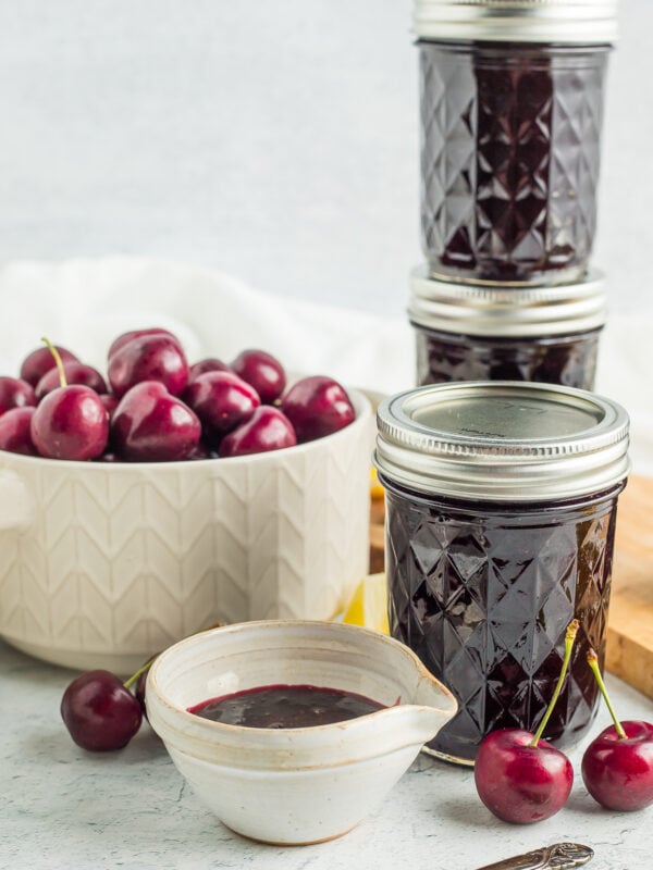three jars and a small dish of cherry preserves with a white bowl of cherries in the background.