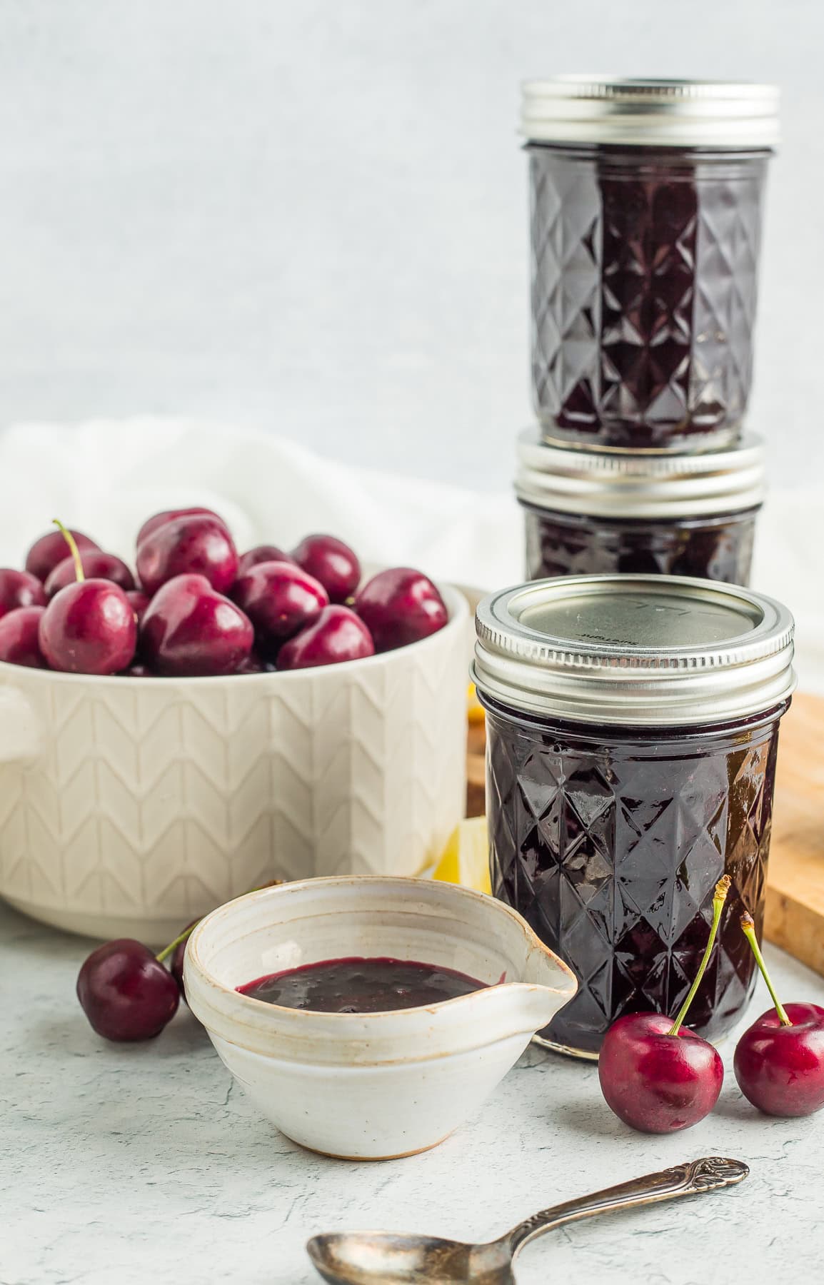 three jars and a small dish of cherry preserves with a white bowl of cherries in the background.