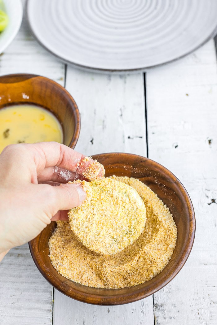 a hand dipping a sliced tomato in breading ingredients.