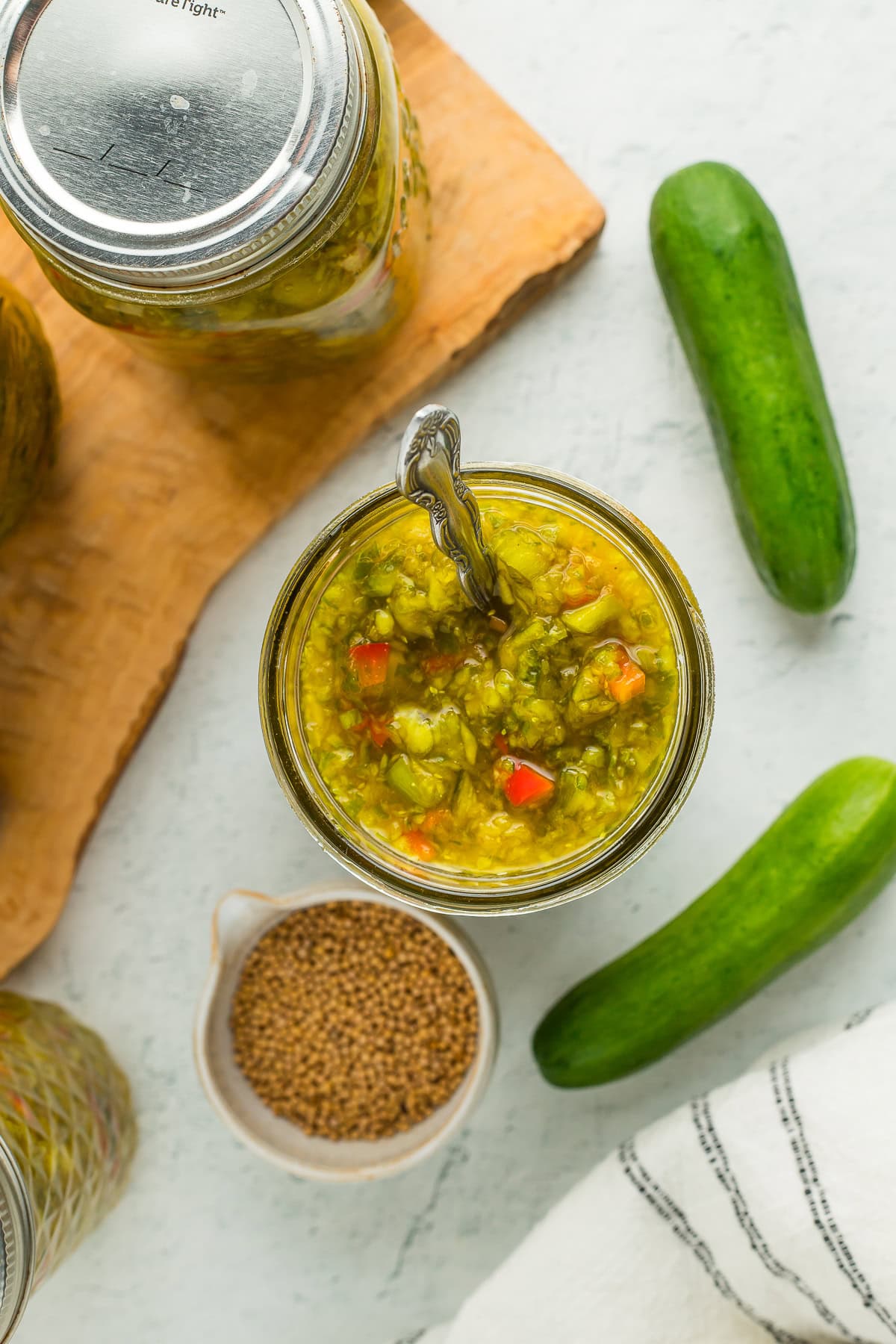 an overhead shot of a jar of canned sweet relish with a spoon in it, a small bowl of mustard seeds, and fresh cucumbers.