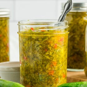 a side profile photo of a canning jar of sweet pickle relish with a small spoon in it, and fresh cucumbers on a white board.