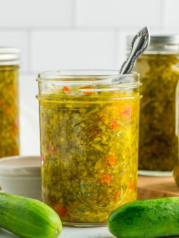 a side profile photo of a canning jar of sweet pickle relish with a small spoon in it, and fresh cucumbers on a white board.