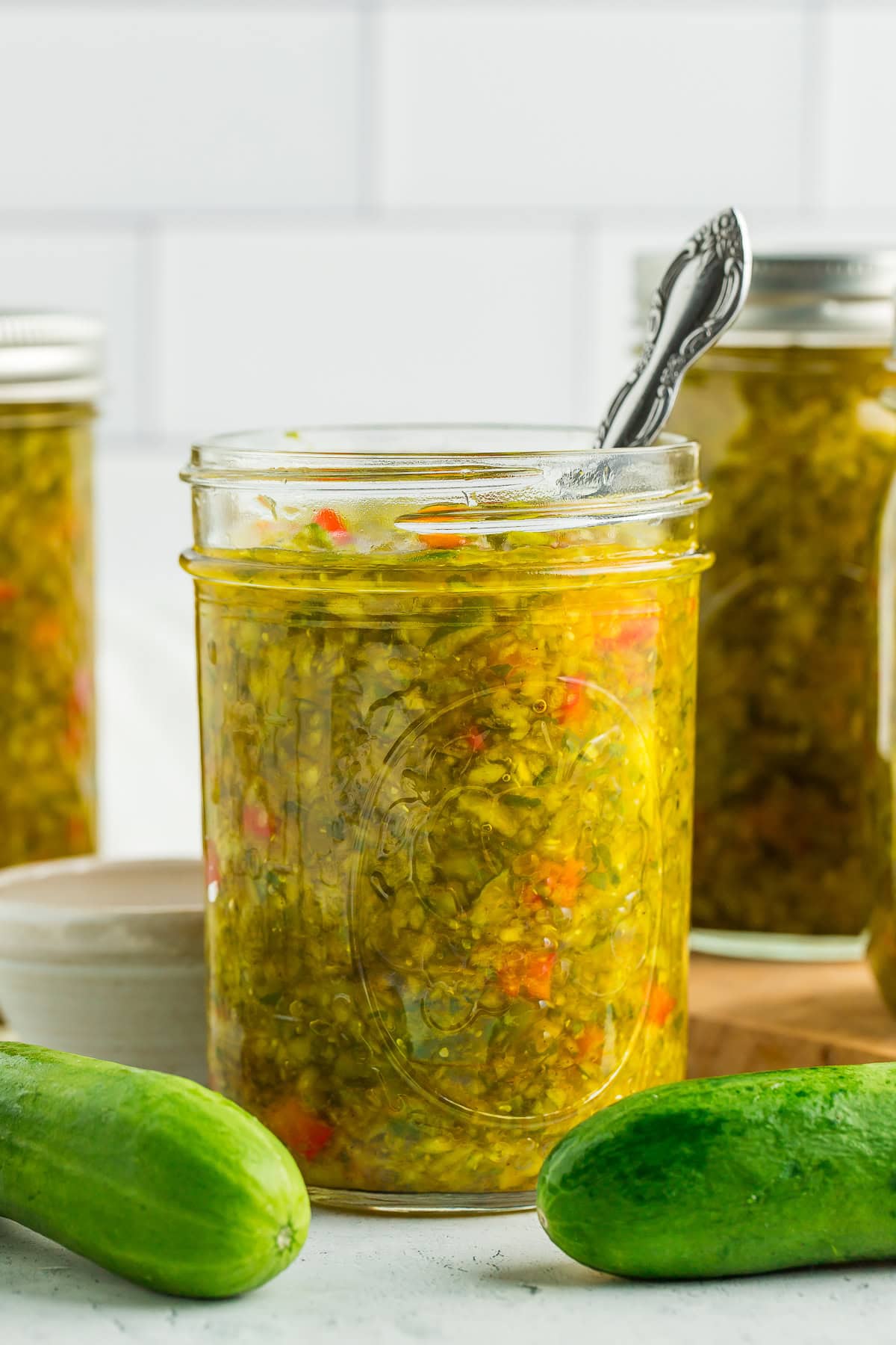 a side profile photo of a canning jar of sweet pickle relish with a small spoon in it, and fresh cucumbers on a white board.