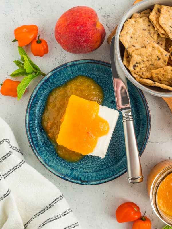 habanero peach preserves on a block of cream cheese on a teal plate with crackers, mint, peaches, and habaneros.