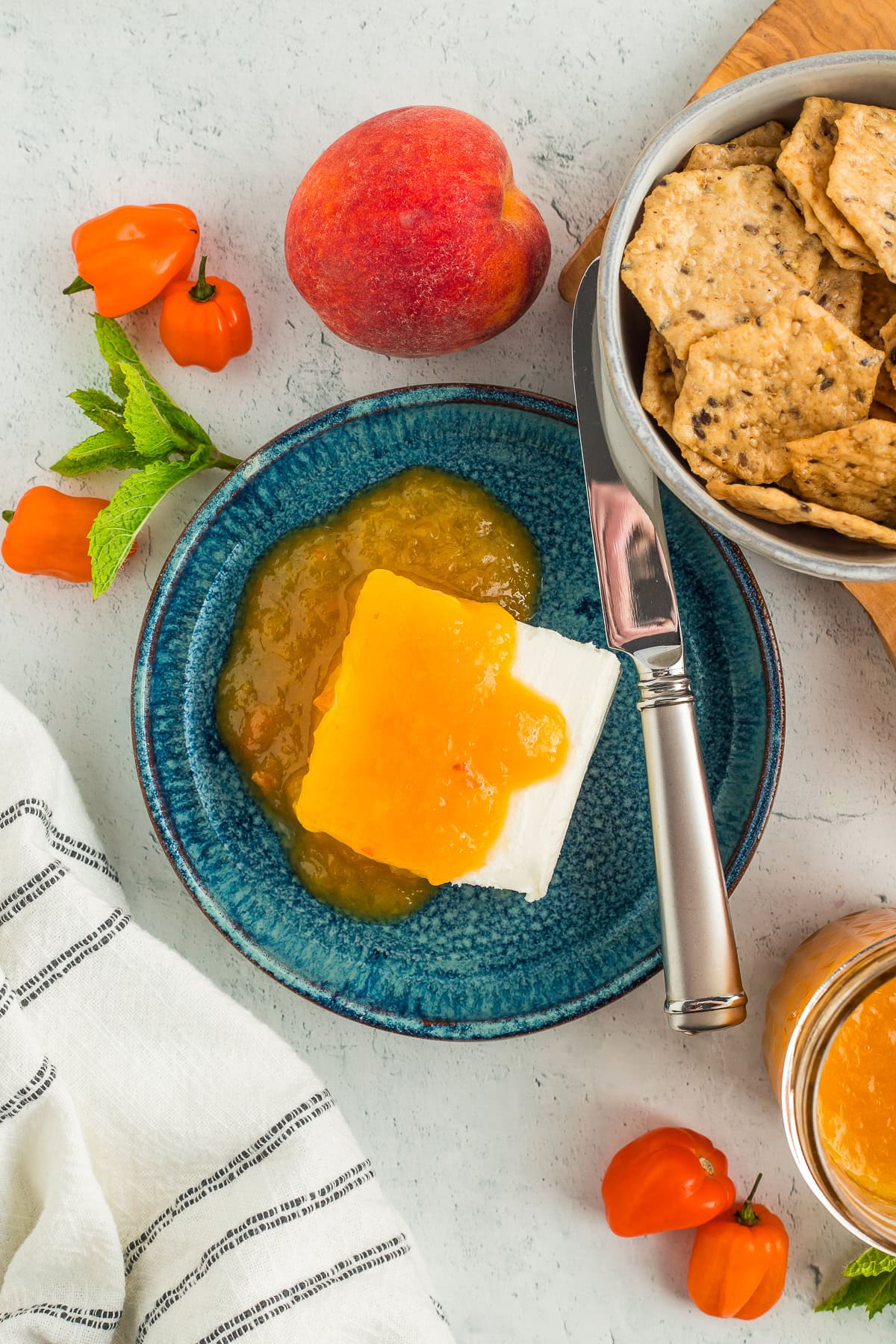 habanero peach preserves on a block of cream cheese on a teal plate with crackers, mint, peaches, and habaneros.