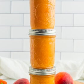 three stacked jars of peach habanero jam with fresh peaches, habaneros, and a spring of mint.