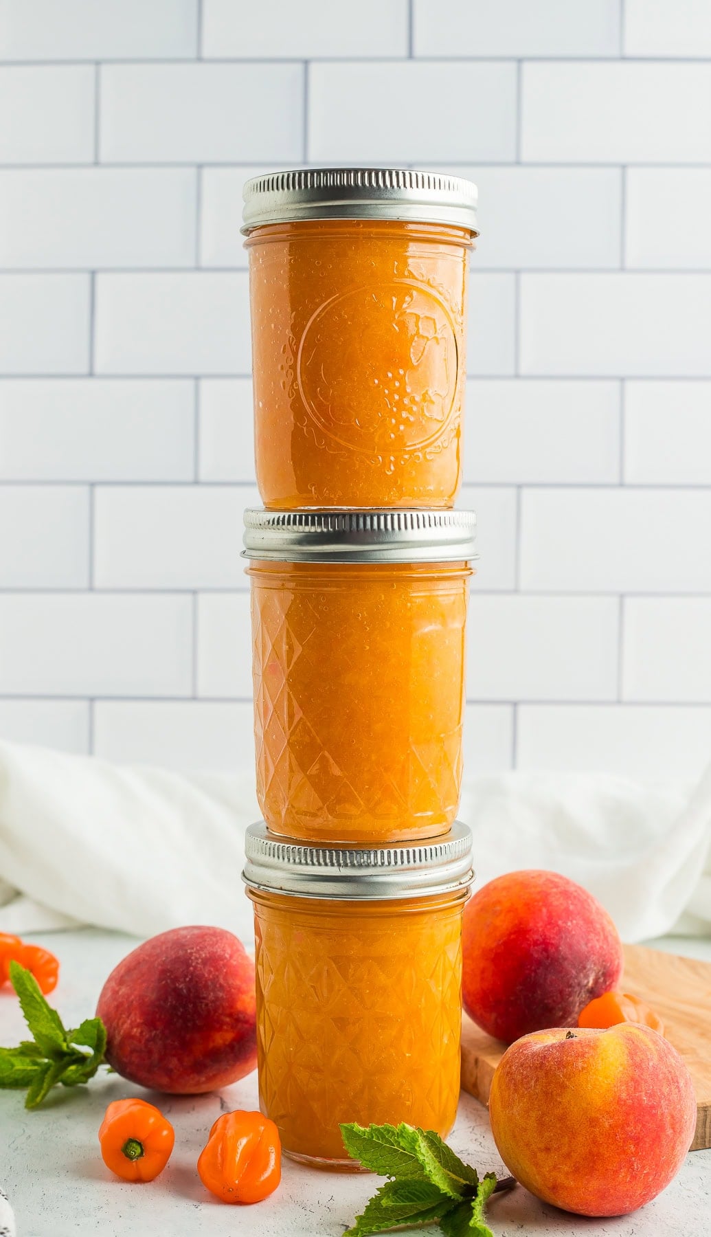 three stacked jars of peach habanero jam with fresh peaches, habaneros, and a spring of mint.