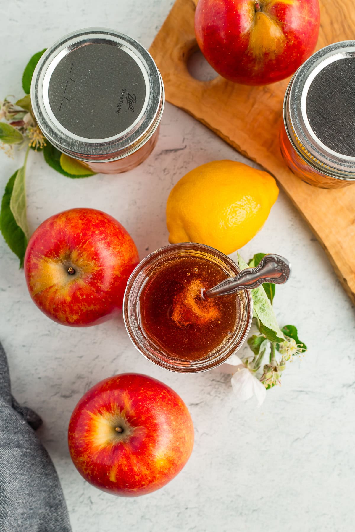 a jar of apple jelly with a spoon in it, fresh apples, and a lemon on a board.