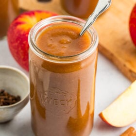 a jar of apple butter with a spoon in it, with cinnamon sticks, apples, and a small bowl of cloves in front of two other jars of apple butter.