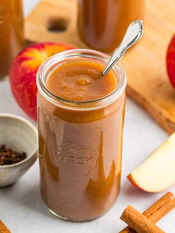 a jar of apple butter with a spoon in it, with cinnamon sticks, apples, and a small bowl of cloves in front of two other jars of apple butter.