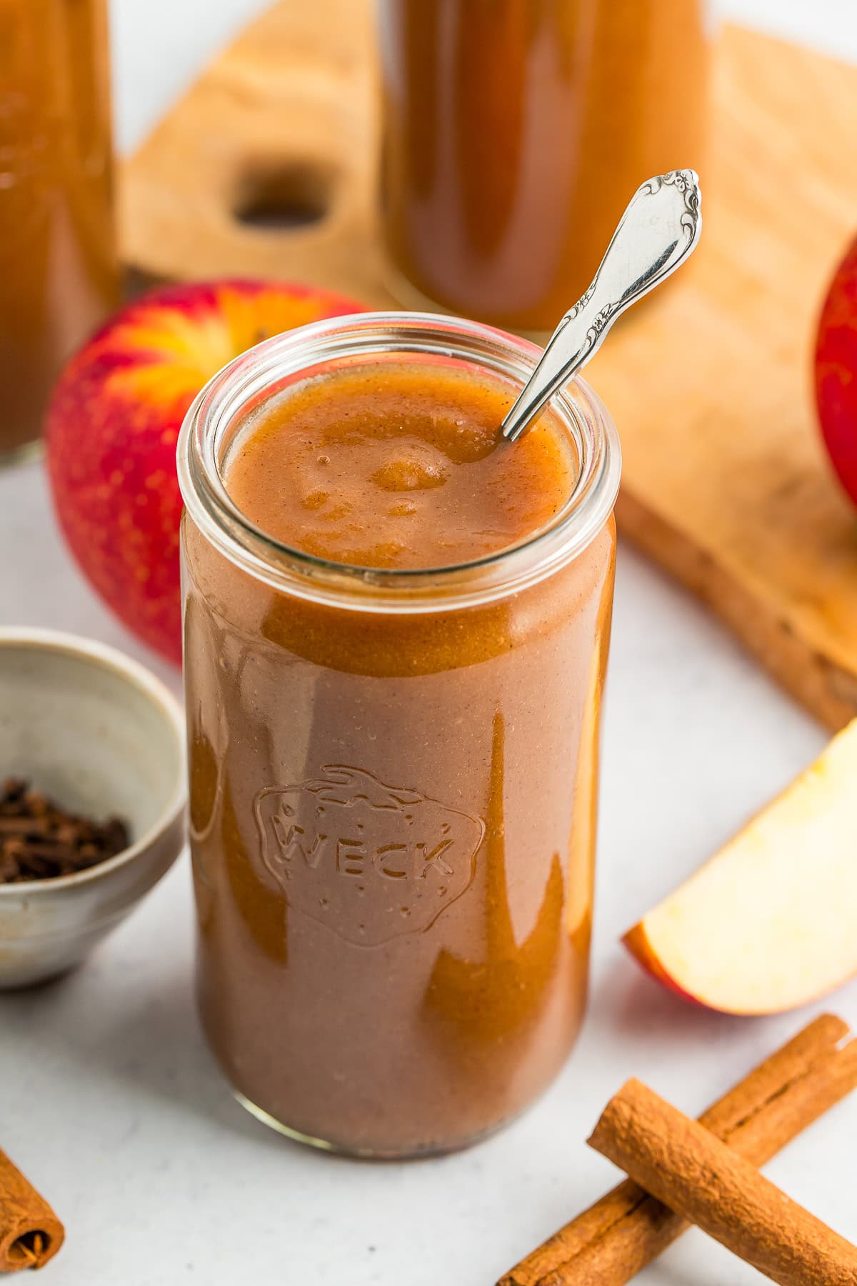 a jar of apple butter with a spoon in it, with cinnamon sticks, apples, and a small bowl of cloves in front of two other jars of apple butter.